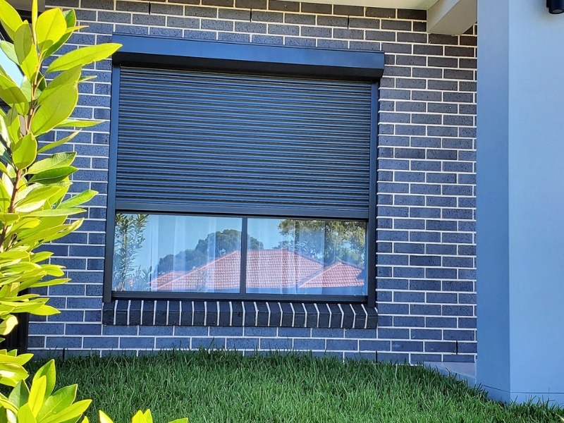 Black roller shutters for temperature control on a modern brick home window, reducing heat and enhancing insulation.