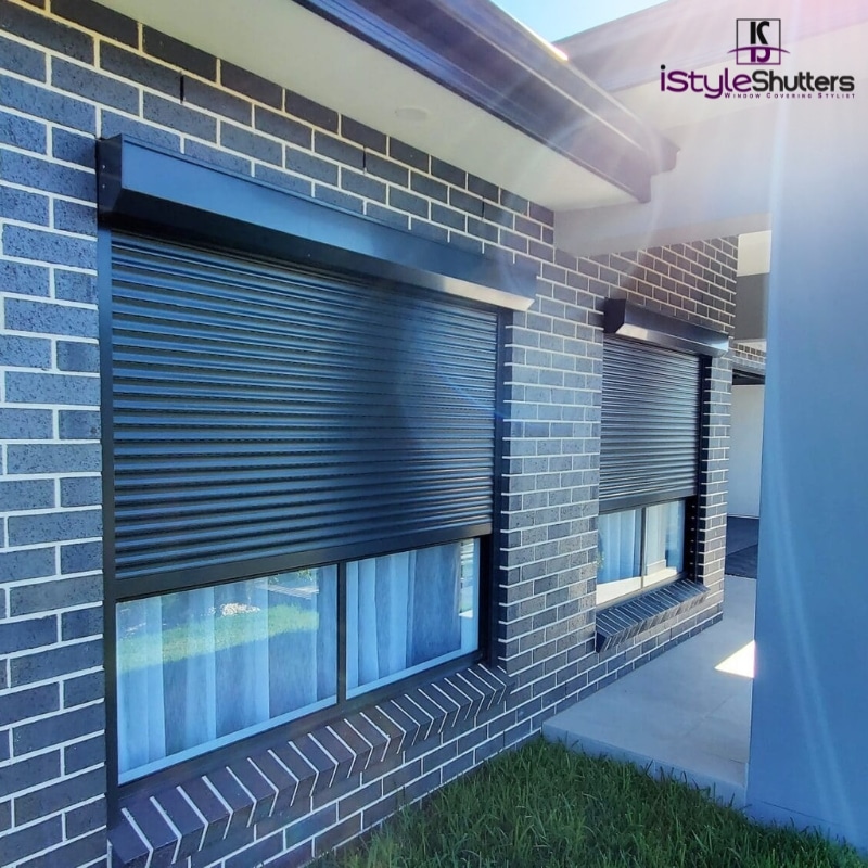 Two sleek black roller shutters in Alfords Point cover the windows of a modern brick home, with soft light filtering through sheer white curtains.