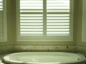 White plantation shutters in coastal homes filter sunlight above a luxurious bathtub.
