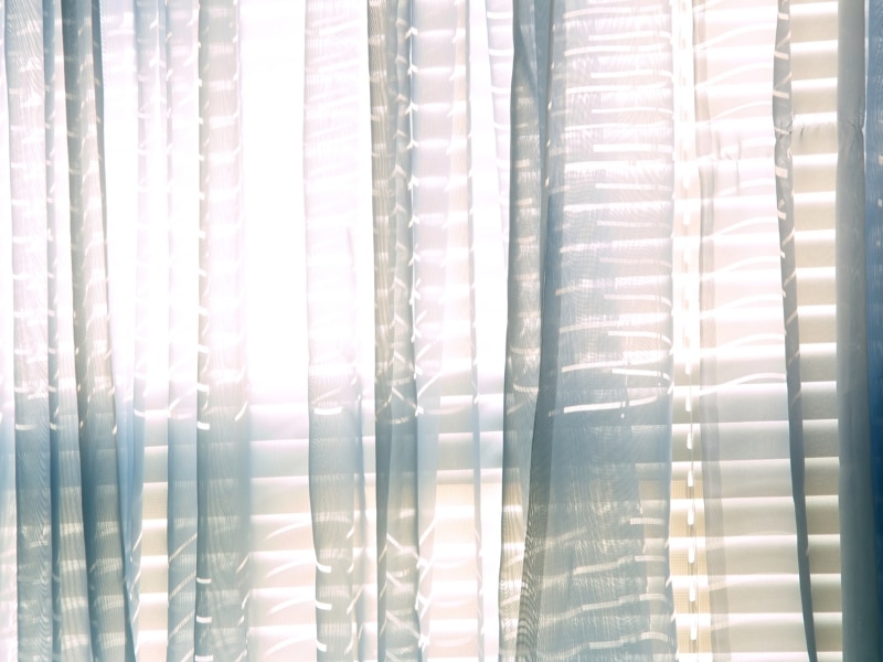 Light streams through a blue sheer curtain over blinds, softly illuminating the window.