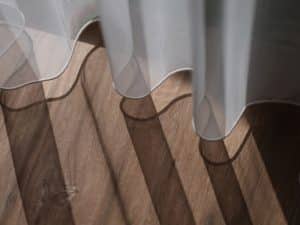 Sunlight streams through a sheer curtain over blinds, casting wavy shadows on the timber floor.