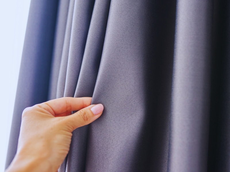 Hand touching thick grey curtain blockout fabric, showcasing its texture and light-blocking quality for home interiors.
