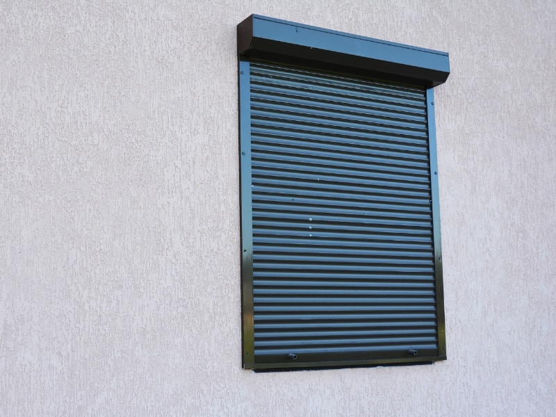 Closed black rolling window shutters installed on an exterior wall of a light-coloured rendered house.