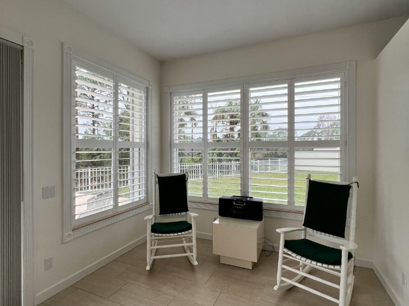 White plantation shutters Sydney installed in a bright living room with two chairs and garden views