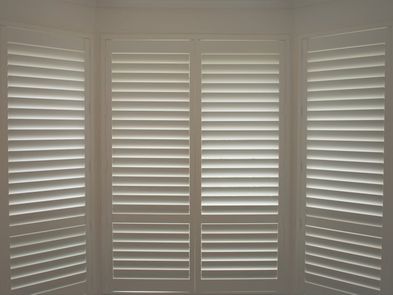 White plantation shutters installed in a bright corner bay window as part of a recent window shutter replacement.